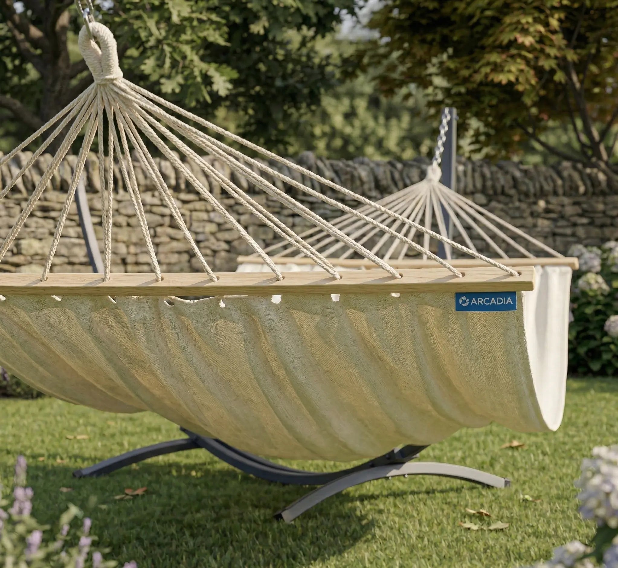 Vue en perspective d'un hamac avec toile crème à barres d'écartement en bois massif monté sur un support en arc anthracite dans un jardin fleuri avec mur en pierre