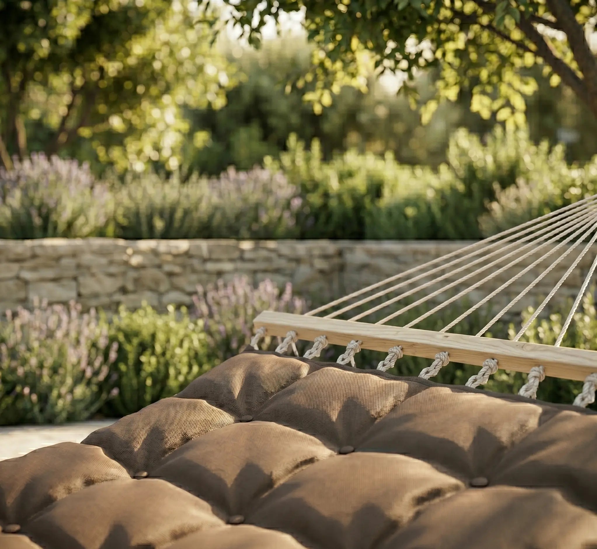 Gros plan sur un hamac matelassé et capitonné de couleur marron avec barre d'écartement en bois, installé dans un jardin luxuriant avec muret en pierre et lavandes en arrière-plan.