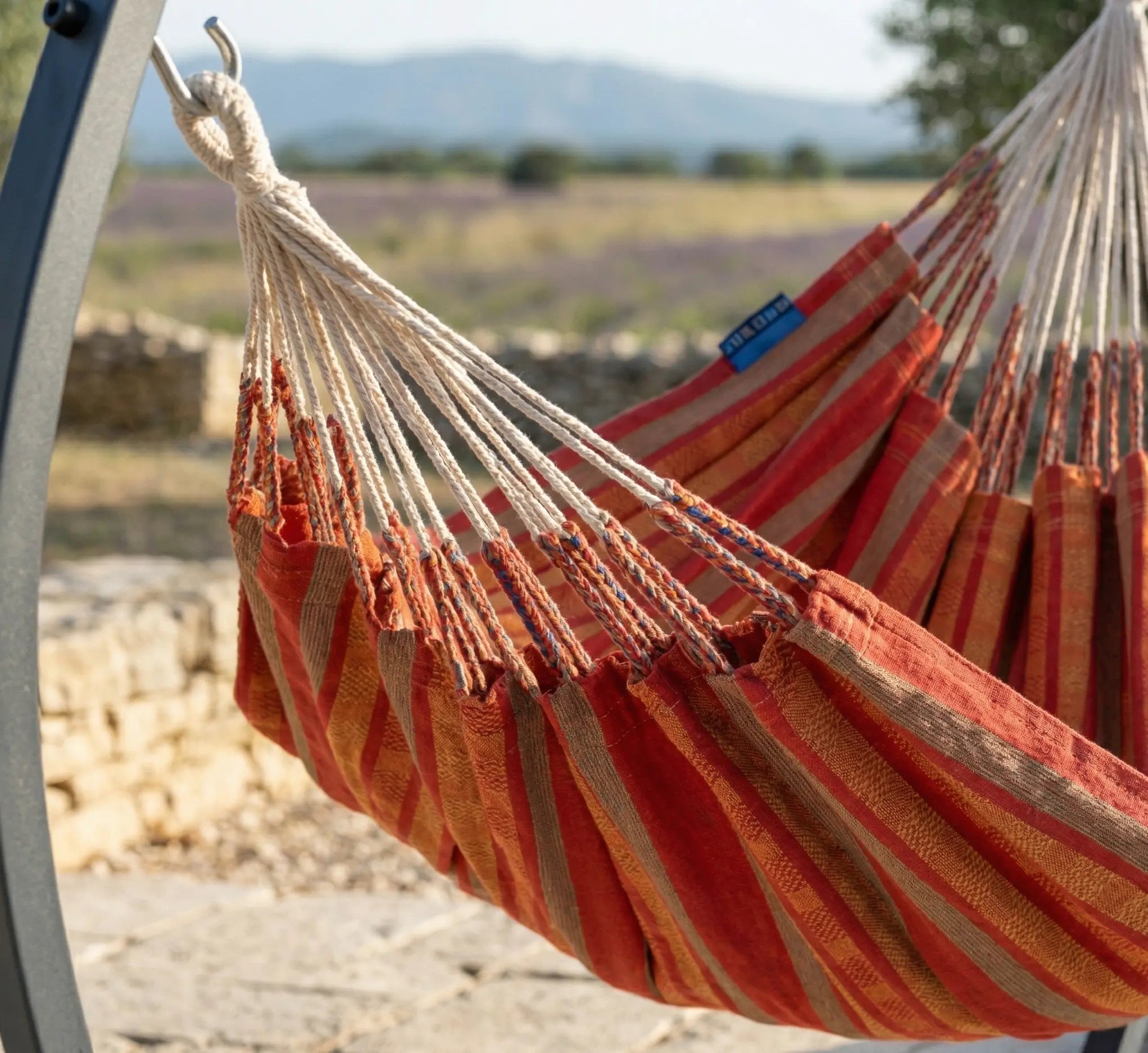 Détail rapproché de la suspension d'une chaise hamac rayée rouge et orange avec de nombreuses cordes de fixation reliées à un crochet en acier robuste sur un support noir en extérieur