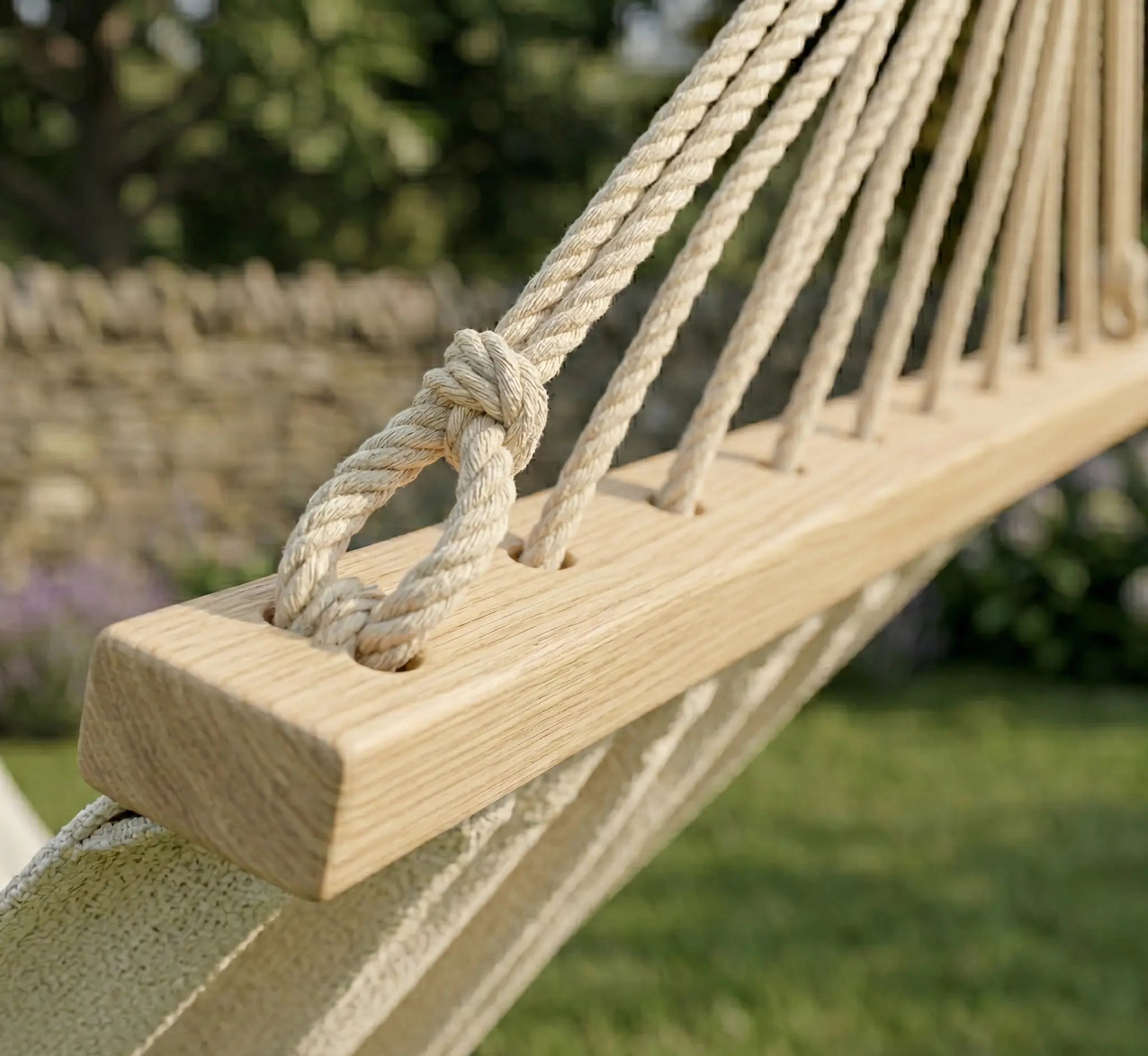 Détail d'une barre d'écartement en bois massif finition chêne avec cordages en coton tressé crème traversant des œillets polis pour un hamac haut de gamme