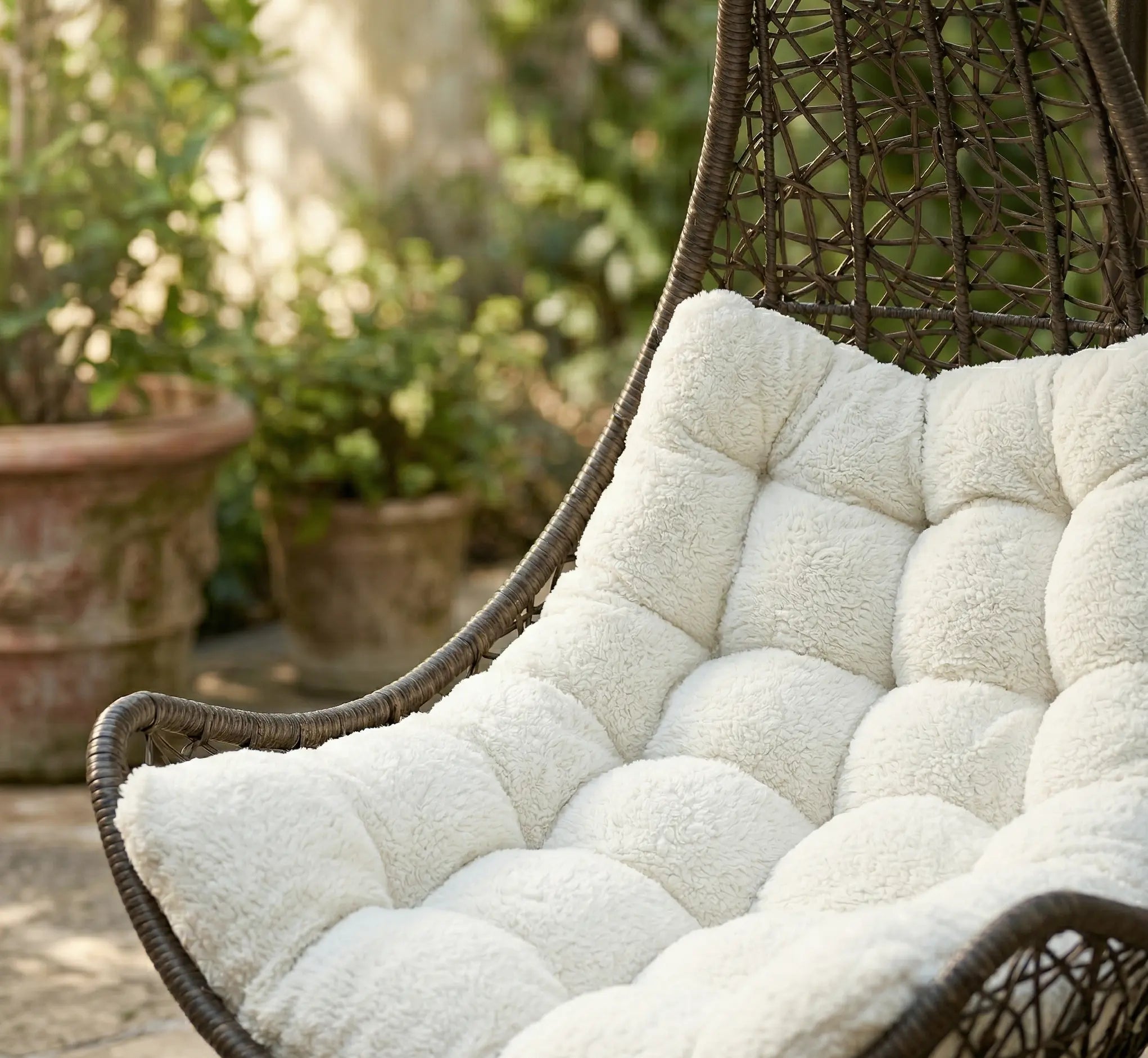 Gros plan sur l'assise d'un fauteuil œuf en résine tressée marron avec un grand coussin blanc capitonné à l'aspect bouclé et douillet dans un jardin ensoleillé