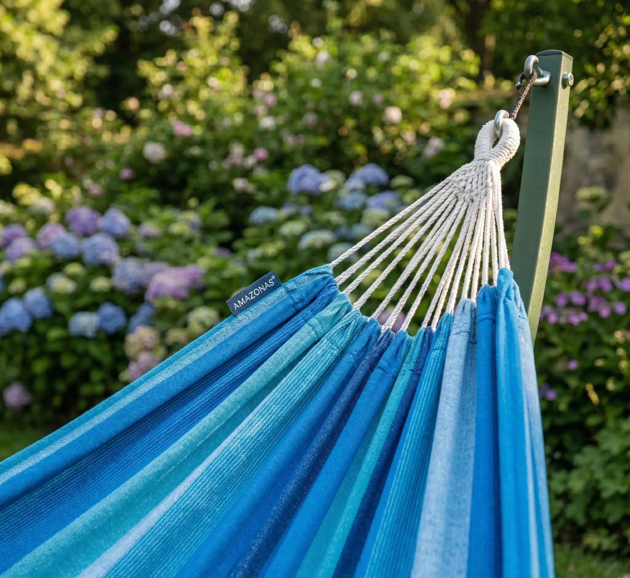 Détail des suspentes blanches tressées d'un hamac aux rayures bleu lagon et bleu ciel montrant la fixation sécurisée sur un montant vert forêt dans un jardin fleuri avec hortensias