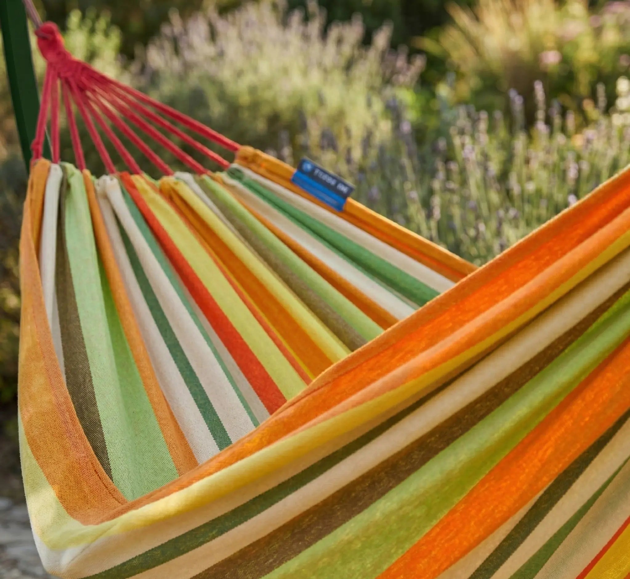 Détail des suspentes tressées blanches d'un hamac aux rayures orange, ocre et jaune montrant la fixation sécurisée sur un montant vert forêt dans un jardin fleuri ensoleillé