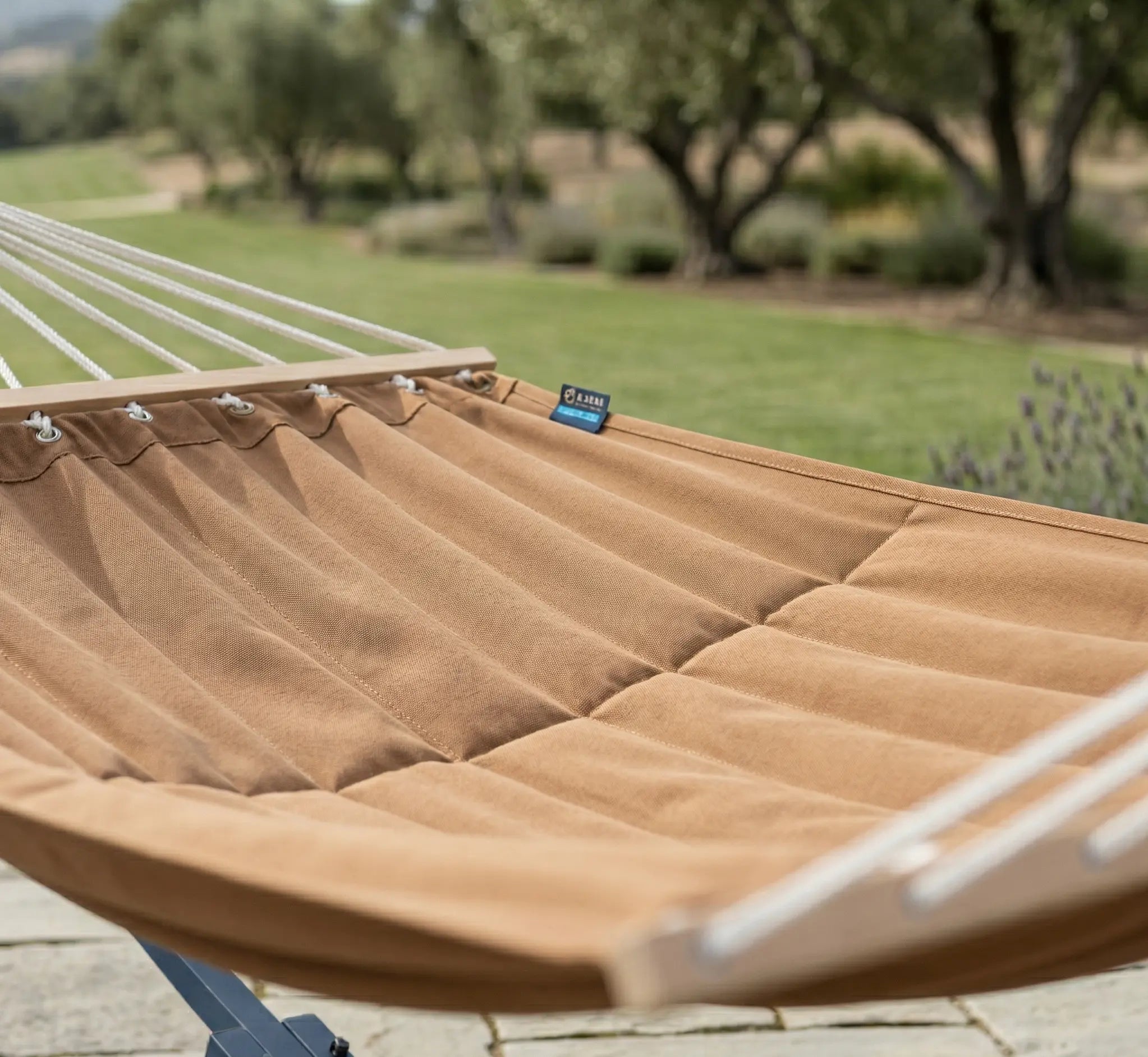 Gros plan sur une toile de hamac moka à l'aspect matelassé avec barres en bois naturel et œillets de fixation, installé sur une terrasse en pierre face à un jardin ombragé